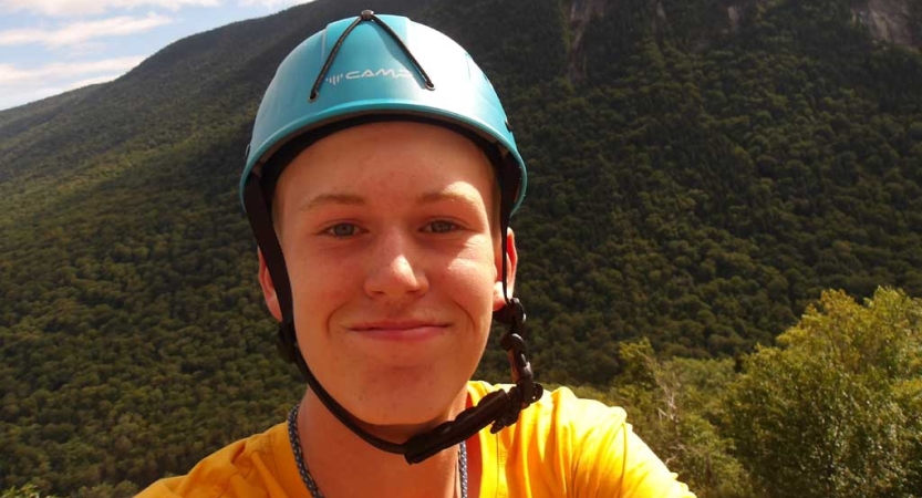 A person wearing a blue helmet smiles at the camera from what appears to be a high elevation. Behind them in the distance is a green slope of trees.. 